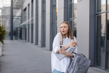 Fototapeta premium Hurry stressed blonde woman in white shirt outside a business center looking anxious as things slip from her hands. She juggles a phone, take away late and book. Bewildered looking girl, confused.