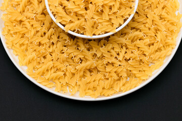 Uncooked Fusilli Pasta Lying on White Plate on Black Background. Raw and Dry Macaroni. Unhealthy and Fat Food - Top View
