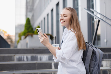 Fototapeta premium Business women style. Woman going to work chatting on phone while going upstairs. Portrait of beautiful smiling female hold take away ice late, reading news on her mobile, look happy, scrolling page.