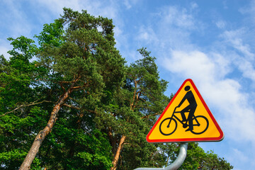 Znak drogowy informacyjny ,, rowerzyści,, na tle błękitnego pochmurnego nieba i wysokich drzew