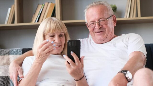 Grandfather and grandmother are sitting at home on the sofa and looking at the smartphone. 