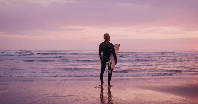 Rear view of a man in a diving suit with wet hair and a surfboard in his hands, standing on the seashore in the pink light of the setting sun and looking into the distance