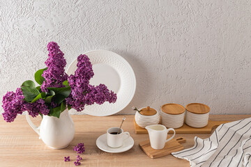 A cup of morning coffee on the kitchen countertop with a bouquet of lilacs in a white jug. Gray cement wall with a copy space. modern kitchen space.