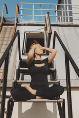 Girl in black sportswear sits in lotus position on stairs on roof, concept of yoga on roofs