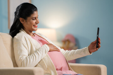Happy indian pregnant woman making video call on mobile phone while sitting on sofa at home -...