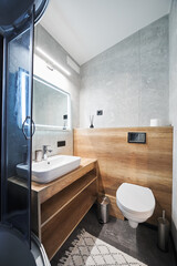 Clean, minimalist grey bathroom in slate with ceramic sink on wooden countertop and hanging toilet inside. Side view of basin and mirror illuminated with warm light in bathroom. Concept of interior.