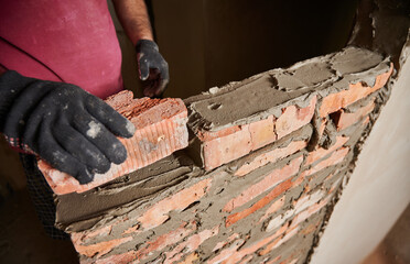 Close up of man hands in work glove laying brickwork in building under construction. Male worker placing brick on block course structure with cement mortar. Masonry construction concept.