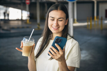 A young woman with coffee talking on the phone in the parking lot.