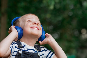 Cute, cheerful, little boy smiles and listens to music in big blue headphones in nature in the...