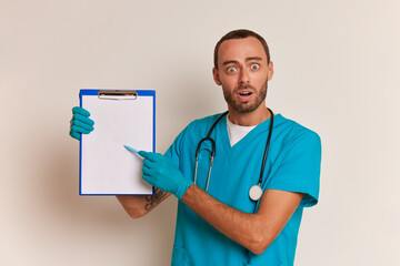 Portrait of young bearded male doctor on white background, wears medical uniform, holds clipboard in hand and asks to sign papers, professional people concept, copy space