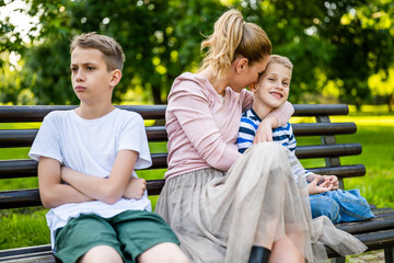 Happy mother is sitting with her sons on bench in park. One boy is offended and pouting. - obrazy, fototapety, plakaty