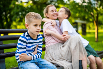 Fototapeta na wymiar Happy mother is sitting with her sons on bench in park. One boy is offended and pouting.