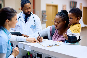 Black mother with son writes data in medical documents while registering in hospital.