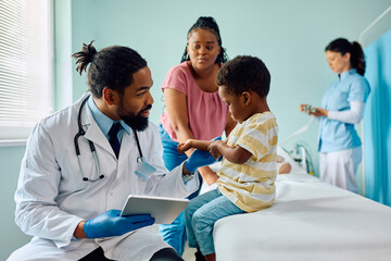 Smiling black pediatrician using touchpad while talking to kid at doctor's office.