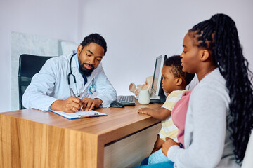 Black pediatrician writing data into medical record during appointment with small boy and his...