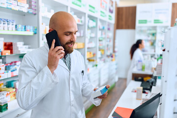 Pharmacist talks on cell phone while holding medicine box in pharmacy.