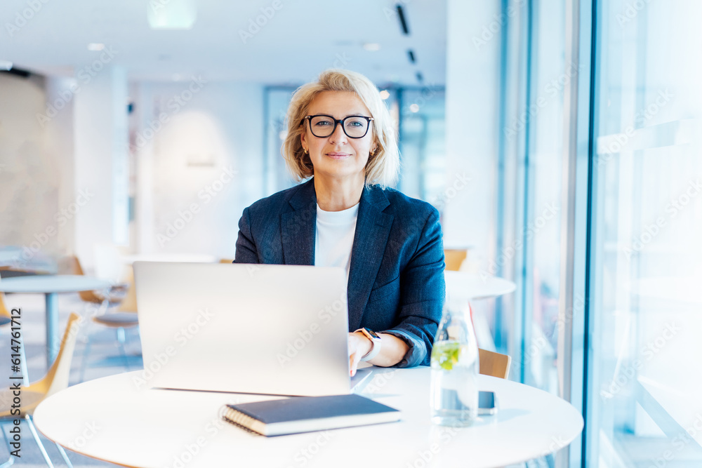 Wall mural Portrait of 50's confident mature businesswoman looking at camera, middle-aged experienced senior female professional working on laptop in open space office. Female entrepreneur working remotely