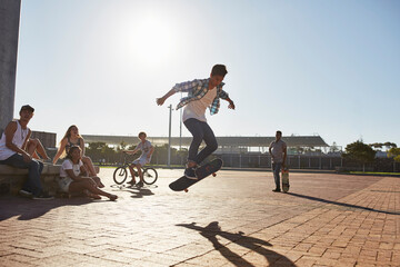 Friends watching teenage boy flipping skateboard at sunny skate park