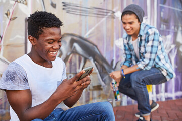 Smiling teenage boys hanging out texting spray painting graffiti on urban wall