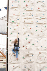 Teenage teen girl grabbing artificial high climbing wall with split ledges. Climbing harness safety...