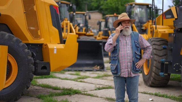 Confident senior man standing on the left talking on phone with yellow huge tractors at background. Portrait of professional Caucasian experienced agrarian choosing new harvester outdoors