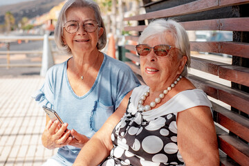 Couple of white-haired senior women sitting on bench in park looking at camera, happy life long friends enjoying retirement