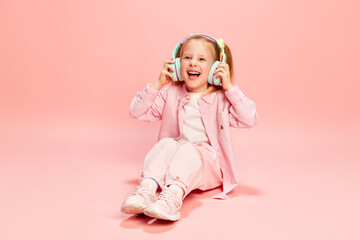 Portrait of happy, cute, beautiful little girl, child listening to music in headphones, smiling against pink studio background