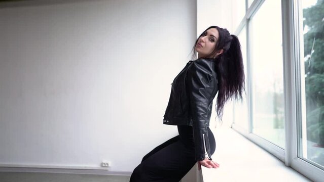 A hot brunette girl in a black dress and a black leather jacket sits on a window in a white studio and looks at the camera
