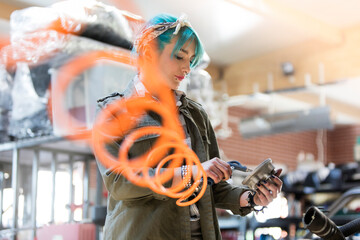 Young female mechanic blue hair using equipment in auto repair shop - obrazy, fototapety, plakaty