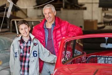 Portrait smiling father son next to classic car in auto repair shop