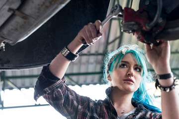 Young female mechanic with blue hair fixing car in auto repair shop - obrazy, fototapety, plakaty