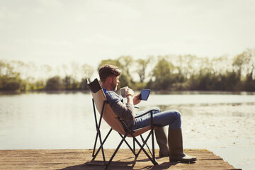 Man drinking coffee and using digital tablet on sunny lakeside dock - obrazy, fototapety, plakaty