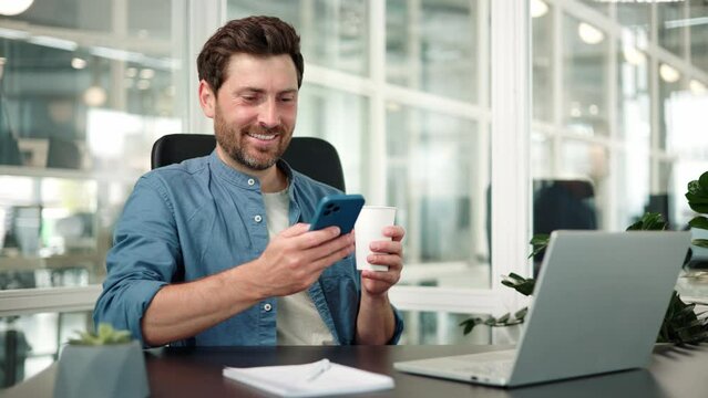Happy businessman reading mobile app notification with coffee in office. Handsome office worker man typing with phone and thinking of ideas for social networking, online connection and internet.