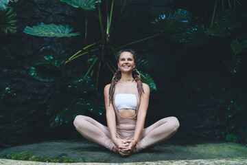 Fototapeta premium Cheerful flexible fitness girl sitting in lotus asana smiling and rejoicing, joyful woman dressed in spandex sportswear recreating on meditation in yoga pose practice for health vitality at nature