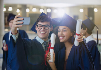 College graduates in cap and gown holding diplomas posing for selfie - obrazy, fototapety, plakaty