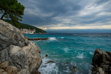 The wave is hitting a rocky seashore. Stormy turqouse blue sea by the promenade in Moscenicka Draga, Croatia. Sea water splash and cloudy sky. Storm and wind. Pine on the rock.