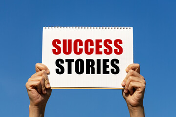Success stories text on notebook paper held by 2 hands with isolated blue sky background. This message can be used as business concept about success stories.
