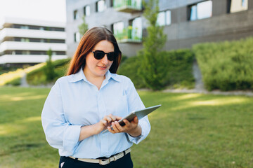 Redhead woman executive smiling confident using touchpad on urban background. Happy young women in a blue shirt and sunglasses walks down the street. Successful business.