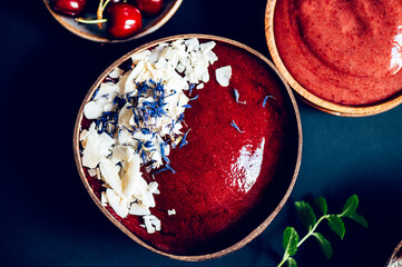 Pink Smoothie from Apple and Cherries with pieces of Coconut Shreds and flower petals on top in the Coconut Shell Bowl with cherries nearby