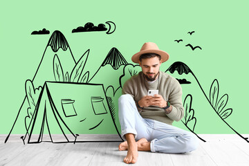 Fototapeta na wymiar Barefooted man with mobile phone sitting near green wall with drawn camping tent and mountains