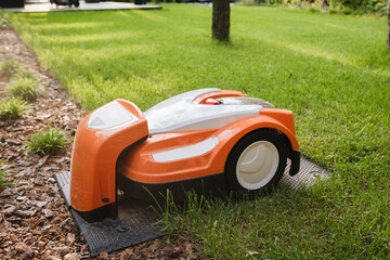 An orange robotic lawn mower stands on the base and is charged from electricity in the courtyard