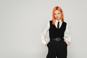 self-assured asian woman with colored red hair posing with hands in pockets in white shirt, black...