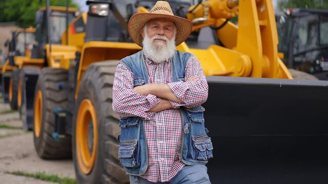 Front view confident senior farmer with crossed hands looking at camera leaning on tractor bucket. Live camera zoom in to satisfied Caucasian bearded old man posing in slow motion outdoors