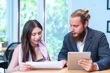 Male hipster bearded market employee holding tablet and female employee sitting together, discussing work information and preparing presentation before presenting at meeting.