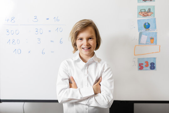 Portrait of cute little happy confident schoolboy smiling stand in classroom with white board math equation on background. Back to school concept. Smart kid boy development