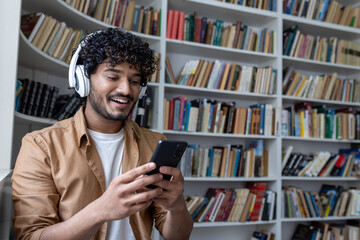 Young hispanic student smiling and happy, using online learning app, smiling man in headphones sitting inside academic library among books.