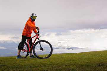 confident cyclist stands with his mountain bike on the edge of a cliff, wearing protective gear. Amazing views of the mountains and nature while doing sports