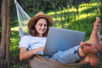 Young cheerful woman working online using laptop lying in hammock in summer garden - 614814326