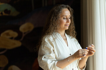 A woman is using rollerball perfume on her wrist. Waist-length portrait by the window, aroma oils and natural fragrances, oil perfumes