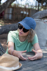 woman explaining and talking in outdoor space with gesticulation on arms, seated at picnic table with cap and heart-shaped sunglasses - obrazy, fototapety, plakaty